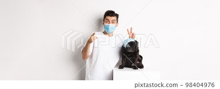 Covid-19, animals and quarantine concept. Happy dog owner and cute pug wearing medical masks, man pointing finger at camera and making funny bunny ears on pet, white background Covid-19, animals and quarantine concept. Happy dog owner and cute pug wearing medical masks, man pointing finger at camera and making funny bunny ears on pet, white background 98404976