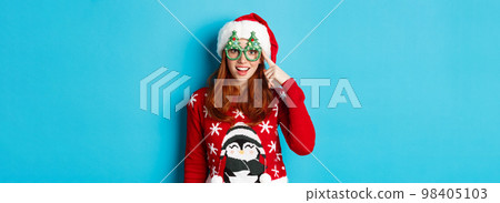 Happy holidays and Christmas concept. Funny redhead teen girl celebrating New Year, wearing santa hat and party glasses, standing against blue background 98405103