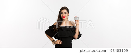 Beauty and shopping concept. Fashionable woman in black dress, wearing makeup, winking at camera and showing credit card, standing over white background Beauty and shopping concept. Fashionable woman in black dress, wearing makeup, winking at camera and showing credit card, standing over white background 98405109