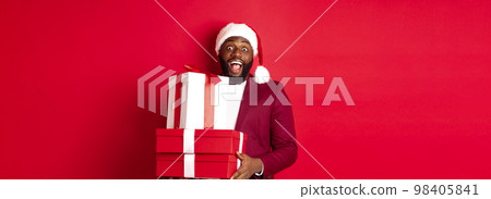 Christmas, New Year and shopping concept. Cheerful Black man secret santa holding xmas presents and smiling excited, bring gifts, standing against red background 98405841
