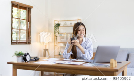 Beautiful Asian woman working on laptop and sipping coffee with smiling face in her home, Creating happiness at work with a smile, Freelancer working at home happily, Work from home. Beautiful Asian woman working on laptop and sipping coffee with smiling face in her home, Creating happiness at work with a smile, Freelancer working at home happily, Work from home. 98405902