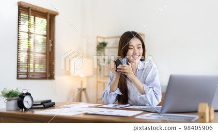 Beautiful Asian woman working on laptop and sipping coffee with smiling face in her home, Creating happiness at work with a smile, Freelancer working at home happily, Work from home. Beautiful Asian woman working on laptop and sipping coffee with smiling face in her home, Creating happiness at work with a smile, Freelancer working at home happily, Work from home. 98405906