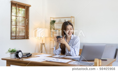 Beautiful Asian woman working on laptop and sipping coffee with smiling face in her home, Creating happiness at work with a smile, Freelancer working at home happily, Work from home. Beautiful Asian woman working on laptop and sipping coffee with smiling face in her home, Creating happiness at work with a smile, Freelancer working at home happily, Work from home. 98405908