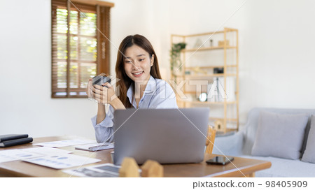 Beautiful Asian woman working on laptop and sipping coffee with smiling face in her home, Creating happiness at work with a smile, Freelancer working at home happily, Work from home. Beautiful Asian woman working on laptop and sipping coffee with smiling face in her home, Creating happiness at work with a smile, Freelancer working at home happily, Work from home. 98405909