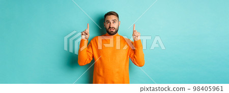 Doubtful and unamused man looking, pointing fingers up something unpleasant, standing skeptical against turquoise background 98405961