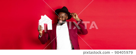 Real estate. Cheerful Black man showing paper house and smiling, recommending broker, standing over red background 98405979