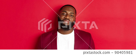 Close-up of handsome bearded plump guy smiling at camera shy, standing in stylish blazer on t-shirt, standing over red background 98405981