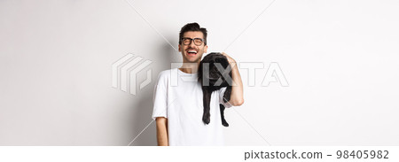 Happy young man laughing carefree, holding cute black dog, pug breed, on shoulder and having fun, standing over white background Happy young man laughing carefree, holding cute black dog, pug breed, on shoulder and having fun, standing over white background 98405982