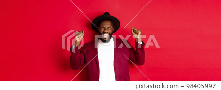Fashion and party concept. Excited Black man winning prize, shaking raised hands and celebrating victory, partying New Year, standing against red background Fashion and party concept. Excited Black man winning prize, shaking raised hands and celebrating victory, partying New Year, standing against red background 98405997