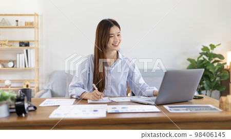 Beautiful young asian woman watching live video or video call of teacher teaching on laptop in her home, Take notes of important conversations and messages during the teacher's teaching. 98406145