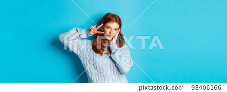 Modern teen girl with red hair, showing peace sign and posing in sweater against blue background Modern teen girl with red hair, showing peace sign and posing in sweater against blue background 98406166