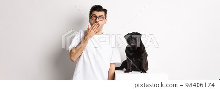 Image of dog owner and cute black pug looking at camera surprised and amazed, standing over white background 98406174