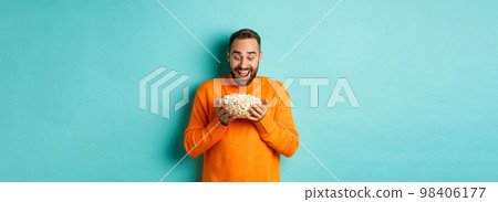 Handsome bearded man looking with exctied and happy at popcorn bowl, ready to watch movie, standing over white background 98406177