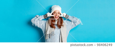 Winter and holidays concept. Cute redhead teen girl in beania, gloves and sweater showing peace sign, looking left at camera and wishing merry christmas, standing against blue background Winter and holidays concept. Cute redhead teen girl in beania, gloves and sweater showing peace sign, looking left at camera and wishing merry christmas, standing against blue background 98406249