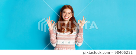 Cute redhead girl in sweater showing okay signs, smiling pleased, approve and like, praise excellent choice, standing over blue background 98406251
