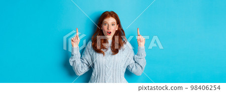 Winter holidays and people concept. Amazed redhead teenage girl pointing fingers up, showing advertisement and staring impressed at camera, blue background 98406254