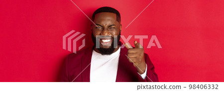 Cheerful Black man with beard wearing party clothes, pointing finger at camera and winking, congratulate with christmas and new year, standing over red background 98406332