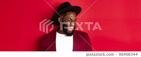 Cheeky african american man in party outfit, winking at camera and smiling, standing against red background 98406344