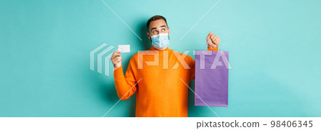 Covid-19, pandemic and lifestyle concept. Thoughtful man in face mask, holding purple shopping bag and credit card, thinking or imaging, standing over turquoise background 98406345