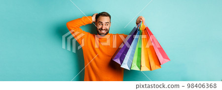 Handsome young man looking awkward and scratching head, staring at shopping bags with gifts, standing over turquoise background Handsome young man looking awkward and scratching head, staring at shopping bags with gifts, standing over turquoise background 98406368