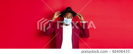 Covid-19, quarantine and holidays concept. Handsome and stylish african american man in face mask, put hat on head and looking sassy, dress-up for party, standing over red background Covid-19, quarantine and holidays concept. Handsome and stylish african american man in face mask, put hat on head and looking sassy, dress-up for party, standing over red background 98406435