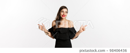 Fashion and beauty. Attractive woman in jewelry, makeup and black dress, smiling and pointing fingers sideways copy space offer, white background Fashion and beauty. Attractive woman in jewelry, makeup and black dress, smiling and pointing fingers sideways copy space offer, white background 98406436
