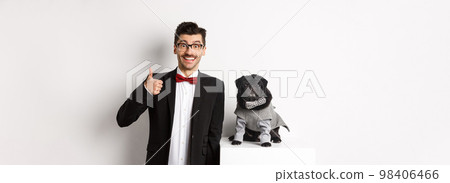 Animals, party and celebration concept. Handsome young man in suit and cute black pug in costume staring at camera, owner showing thumb up in approval and praise, white background 98406466