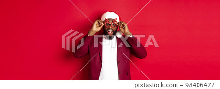 Funny Black man celebrating New Year, wearing party glasses and santa hat, having fun over red background 98406472