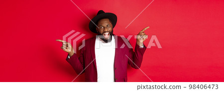 Winter holidays and shopping concept. Cheerful Black man smiling and showing two promo, pointing fingers sideways at copy spaces, red background Winter holidays and shopping concept. Cheerful Black man smiling and showing two promo, pointing fingers sideways at copy spaces, red background 98406473
