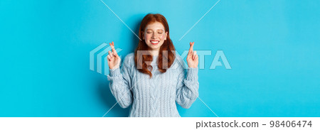 Hopeful redhead girl making a wish, cross fingers for good luck, smiling and anticipating good news or positive result, standing against blue background Hopeful redhead girl making a wish, cross fingers for good luck, smiling and anticipating good news or positive result, standing against blue background 98406474