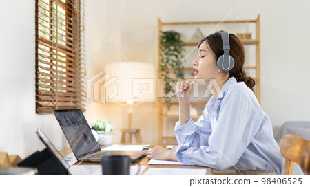 Asian female college student wearing headphones watching live performance or video call teacher teaching on laptop, Conversations with teachers and classmates, Online learning, Study at your own home. 98406525