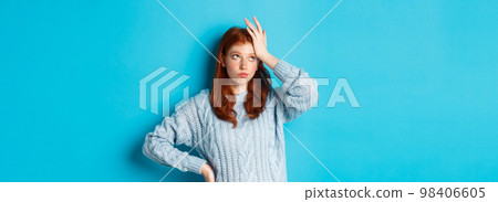 Annoyed and tired redhead teenage girl roll eyes, facepalm and sighing bothered, standing in sweater against blue background 98406605