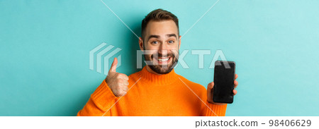 Close-up of attractive bearded man showing smartphone screen, thumbs up, recommending mobile app, standing satisfied over turquoise background Close-up of attractive bearded man showing smartphone screen, thumbs up, recommending mobile app, standing satisfied over turquoise background 98406629