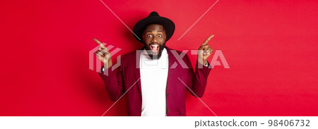 Winter holidays and shopping concept. Cheerful Black man smiling and showing two promo, pointing fingers sideways at copy spaces, red background Winter holidays and shopping concept. Cheerful Black man smiling and showing two promo, pointing fingers sideways at copy spaces, red background 98406732