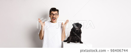 Cheerful young man in glasses standing with his pet, rejoicing and staring at camera amused, hear great news, standing with pug over white background Cheerful young man in glasses standing with his pet, rejoicing and staring at camera amused, hear great news, standing with pug over white background 98406744