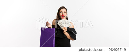 Image of excited beautiful woman in black dress going shopping, holding bag and dollars, standing over white background 98406749