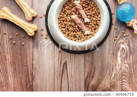 A bowl with dog food, dog treats and toys on a wooden floor. 98406759