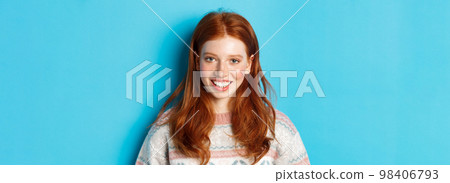 Close-up of cute redhead girl in sweater smiling happy at camera, standing against blue background Close-up of cute redhead girl in sweater smiling happy at camera, standing against blue background 98406793