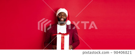 Christmas, New Year and shopping concept. Happy bearded african american man holding xmas present, smiling at camera, standing in santa hat against red background 98406821