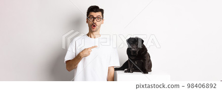 Happy young man showing his cute dog, pointing finger at black pug and staring amazed, standing over white background 98406892