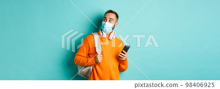 Young man in face mask using mobile phone, holding backpack, staring right amazed, standing against light blue background Young man in face mask using mobile phone, holding backpack, staring right amazed, standing against light blue background 98406925
