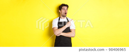 Waiter in black apron checking out promo offer, pointing finger and looking left at your logo, standing over yellow background 98406965