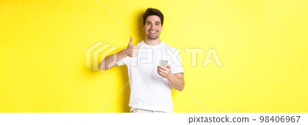 Happy satisfied man holding smartphone, showing thumb up in approval, recommend something online, standing over yellow background 98406967