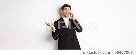 Portrait of happy good-looking businessman receiving great offer, talking on phone and looking pleased, standing in black suit against white background 98407008