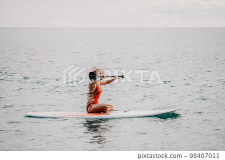 Woman in red bikini on sup board. Happy lady with blond hair in red bathing suit chilling and sunbathing by turquoise sea ocean on sunset. Holiday, vacation and recreational concept. Woman in red bikini on sup board. Happy lady with blond hair in red bathing suit chilling and sunbathing by turquoise sea ocean on sunset. Holiday, vacation and recreational concept. 98407011