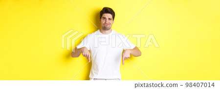 Young unhappy guy grimacing, pointing fingers down at advertisement, disappointed in product, standing over yellow background Young unhappy guy grimacing, pointing fingers down at advertisement, disappointed in product, standing over yellow background 98407014