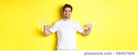 Happy attractive guy pointing fingers at your logo, showing promo on his t-shirt, standing over yellow background 98407404