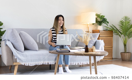 Woman using laptop to work or do homework at home with smiling face in her living room, Creating happiness at work with a smile, Live performance or video call with laptop, Work from home. Woman using laptop to work or do homework at home with smiling face in her living room, Creating happiness at work with a smile, Live performance or video call with laptop, Work from home. 98407414