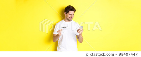 Young man paying online, insert credit card number on mobile phone, shopping in internet, standing over yellow background 98407447