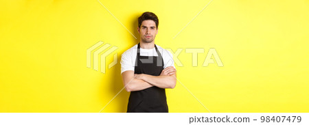 Suspicious barista squinting, cross arms on chest and looking at something with disbelief, standing over yellow background Suspicious barista squinting, cross arms on chest and looking at something with disbelief, standing over yellow background 98407479